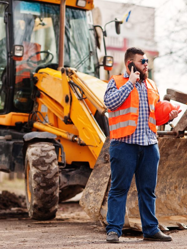 Beard worker man suit construction worker in safety orange helmet, sunglasses against tractor with mobile phone at hand.